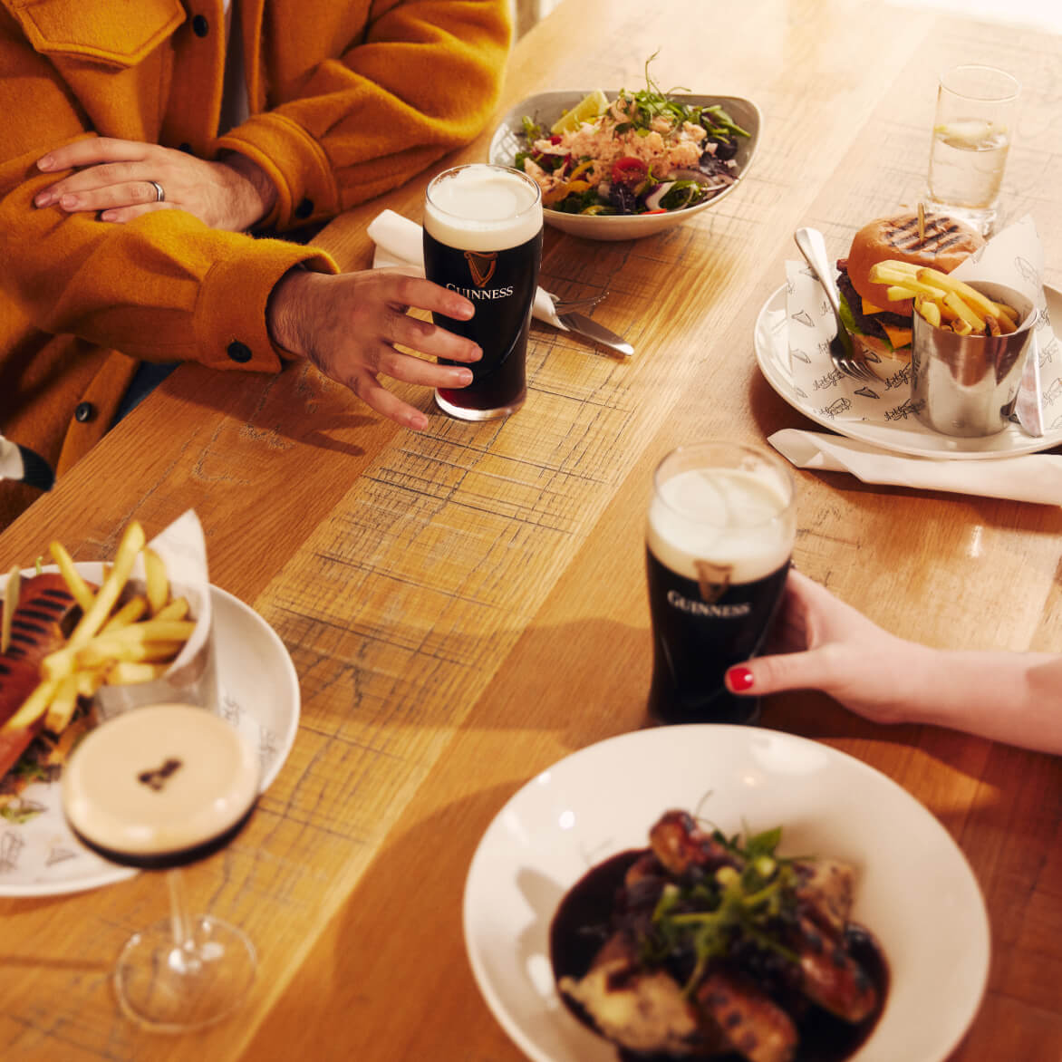 Table with 2 beautiful pints of smooth Guinness, a glass of chilled tap water, and a foamy espresso martini along with a succulent burger and chips, bangers and mash covered in a delicious homemade gravy, a fresh salad, and a tasty steak sandwich and chips. 