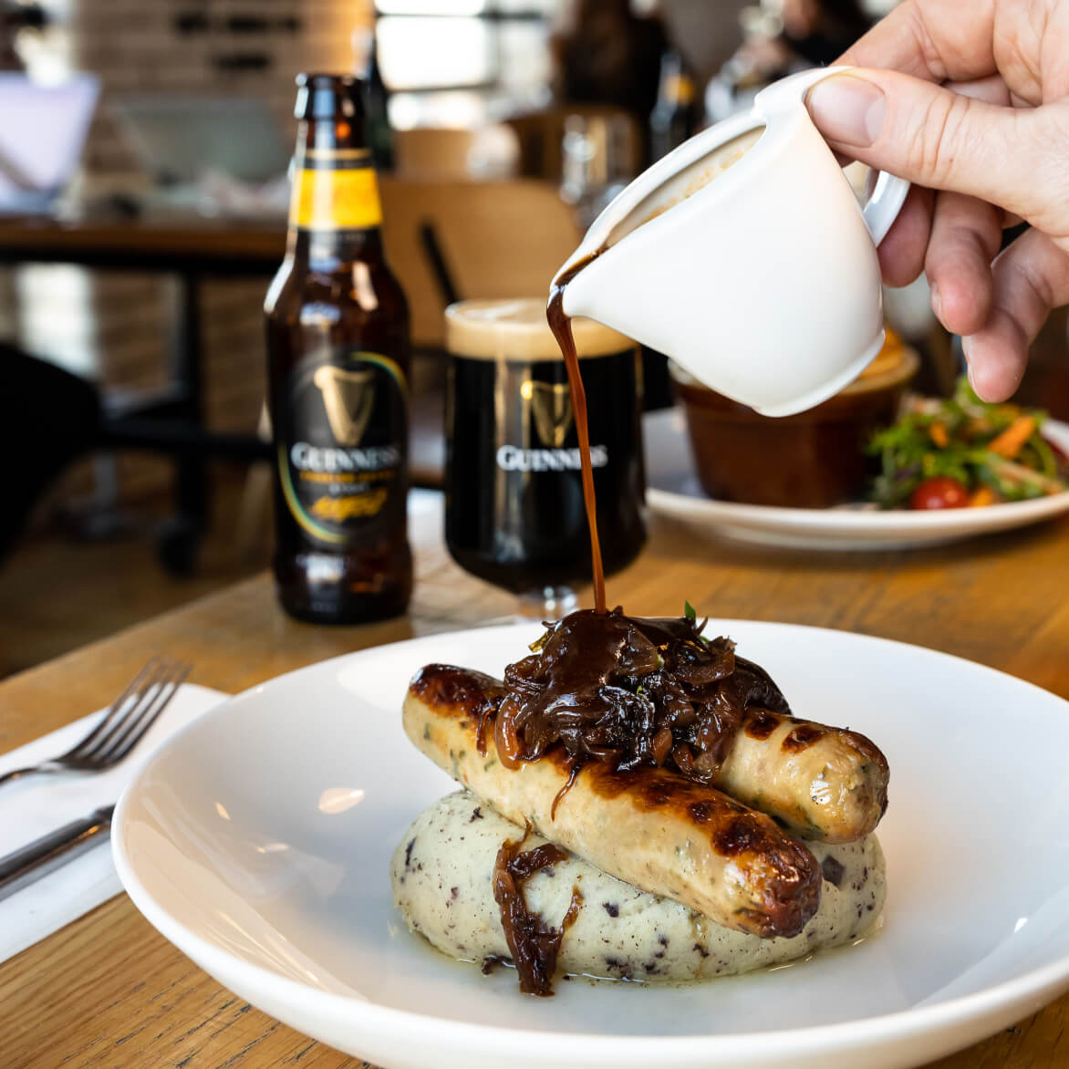 Someone pouring Guinness & red onion gravy over their bangers & mash, recommended to be served with Guinness foreign extra stout which can be seen blurred in the background.