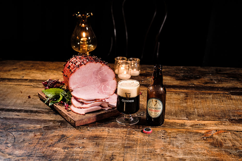 A Guinness cured ham on a wooden table with a glass of Guinness original beside it