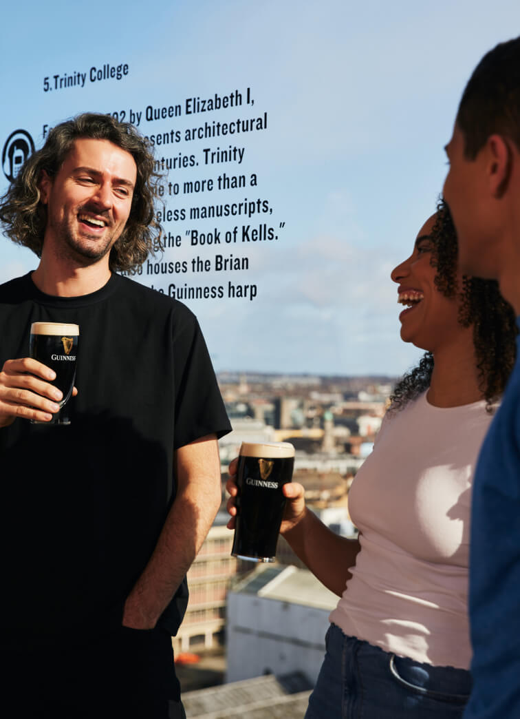 Two men and one woman smiling and chatting while holding perfectly poured pints of creamy Guinness. The background is a breath-taking view of Dublin city and specifically Trinity College as can be seen above the man’s curly hair. 