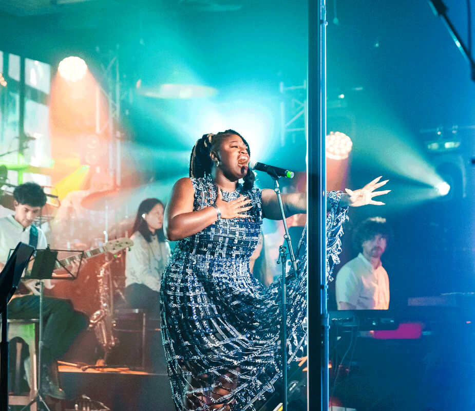 A woman in a blue sparkly dress sings on stage with her arm outstretched, orchestra around her