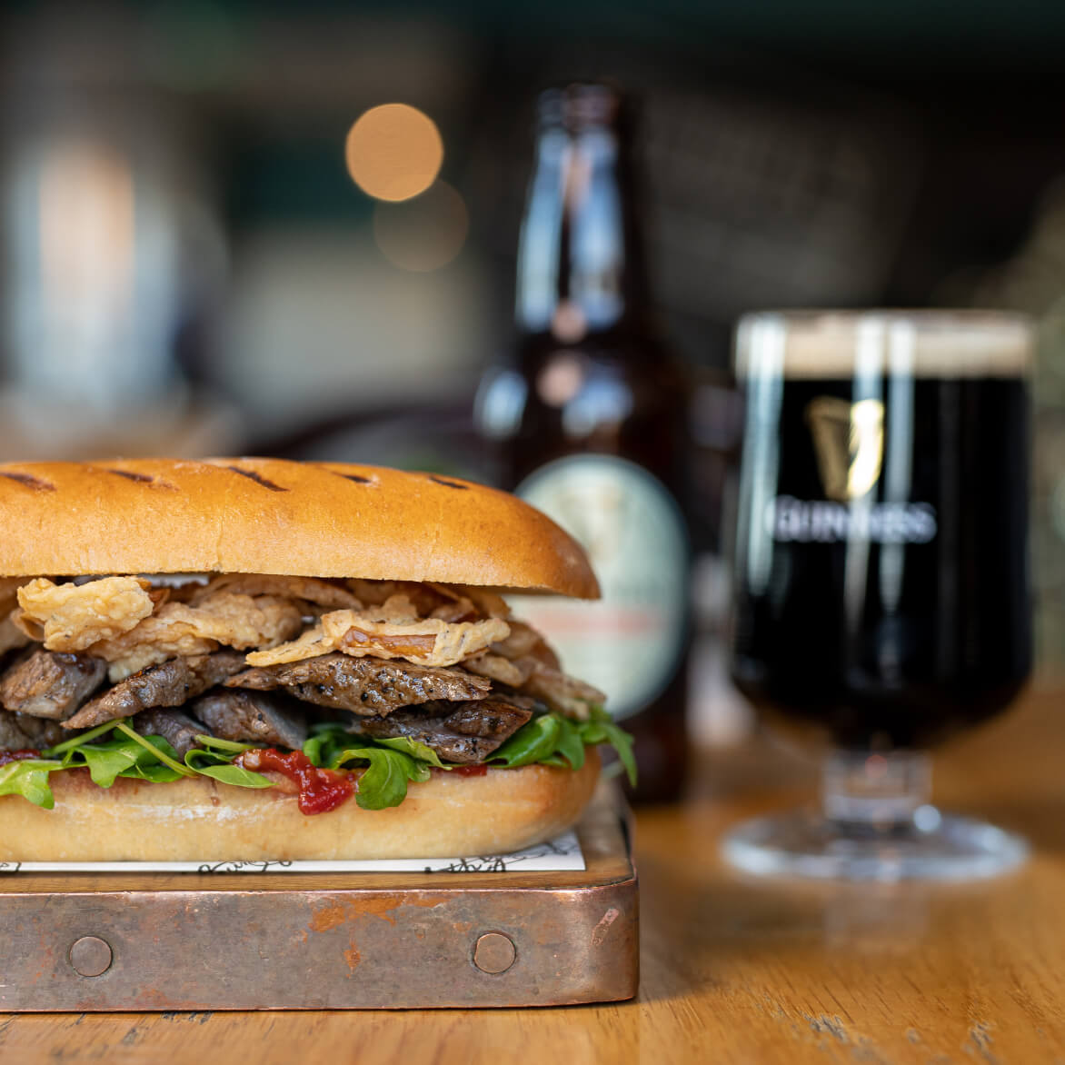 Arthur’s Bar steak sandwich made from prime Irish striploin, served on a brioche sub roll, with spicy Ballymaloe relish and crispy onions. Guinness Original Extra Stout can be seen behind the sandwich which is the recommended beer pairing. 