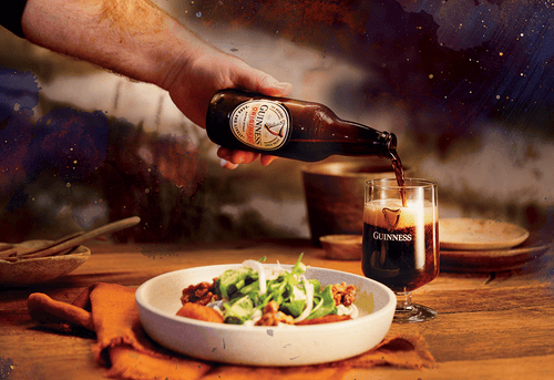 Man's hand pouring Guinness Original into glass on a table, beside a plate of butternut squash fritters