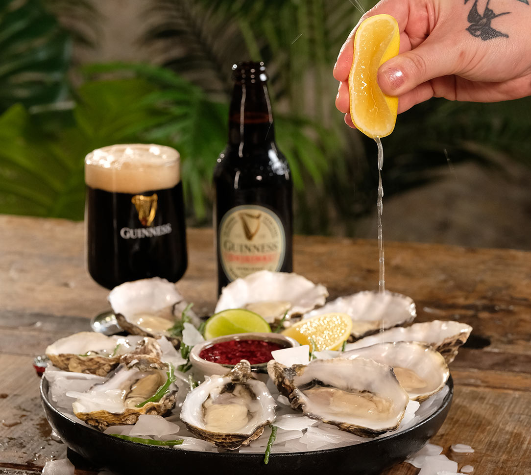 A hand squeezing a lemon over a plate of oysters, glass and bottle of Guinness in the background