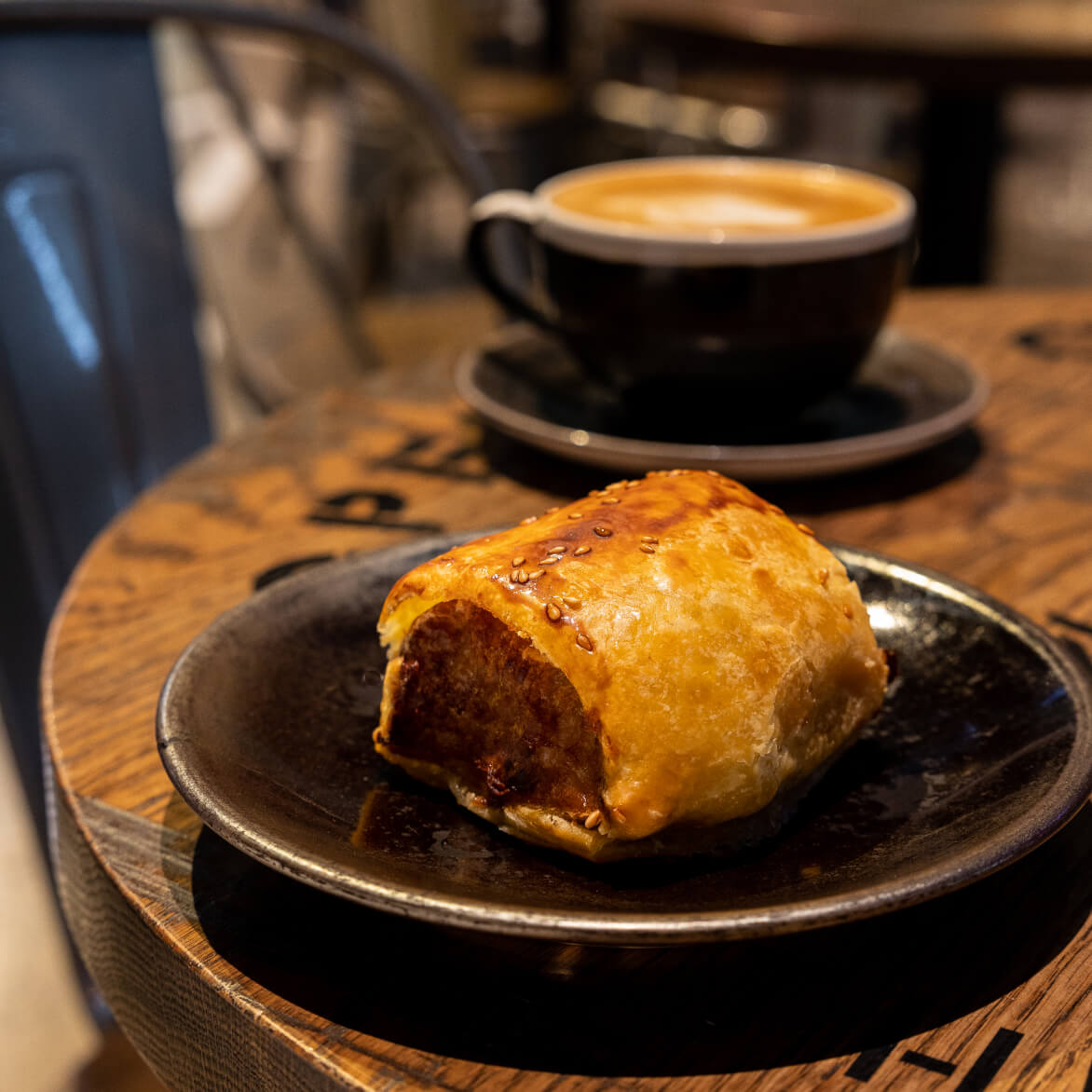 A fresh sausage roll available at the Cooperage Café with a blurred coffee in the background.