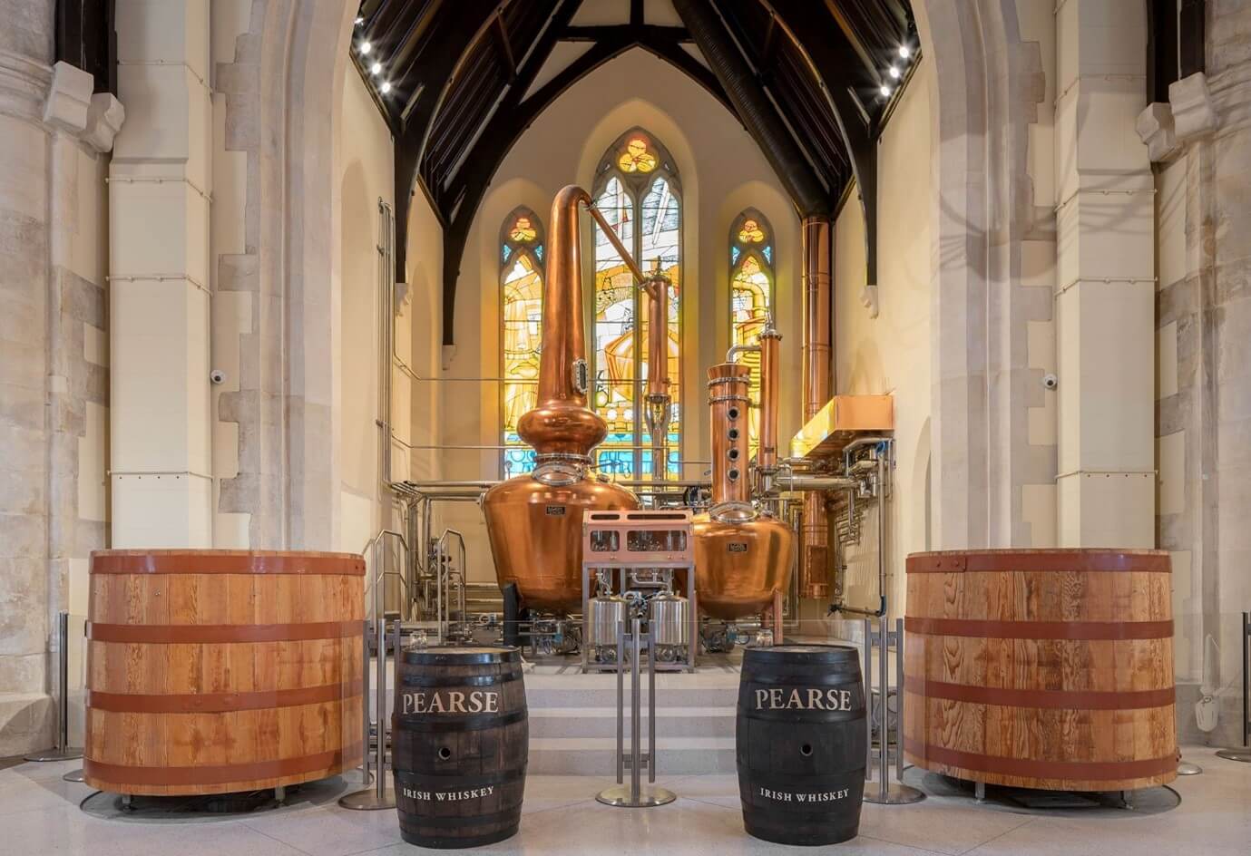 The inside of Pearse Lyon Distillery. Small and Large whiskey barrels can be seen with a large whiskey pot still in the back of the room. There is painted stained glass windows reflecting in vibrant lighting.