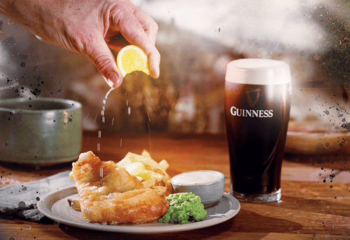 A hand squeezes lemon over a plate of fish and chips, beside a pint of Guinness, all on a wooden table