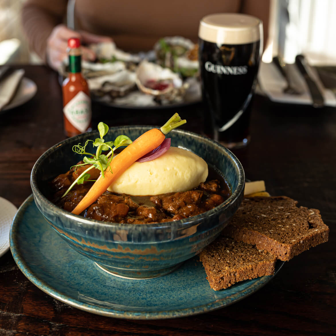 Bowl of signature beef stew with mash potato, baby carrot, two slices of soda bread on the side, and a pint of Guinness.