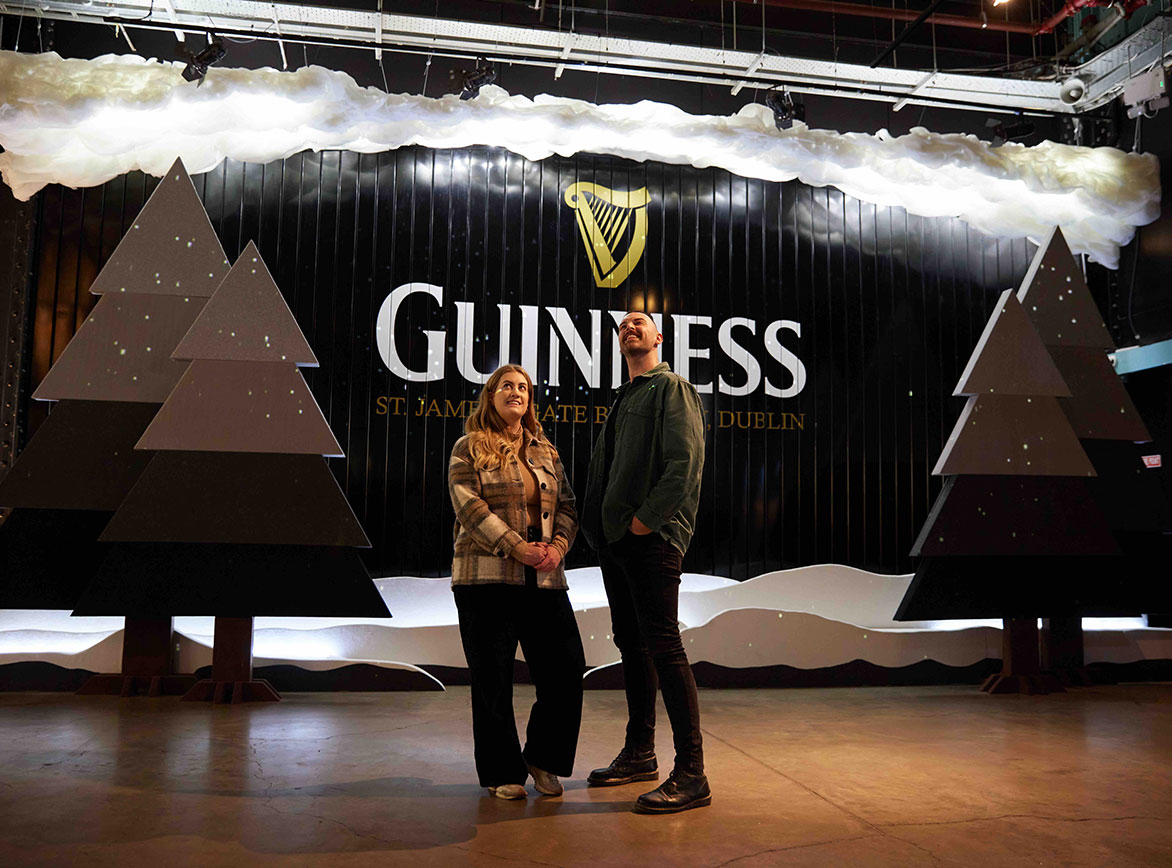 A man and woman stand in front of a decorated Guinness gate, with large grey Christmas trees beside them