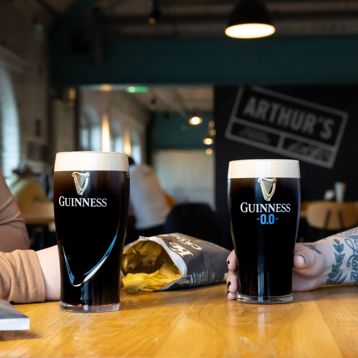 Two people talking over a Guinness draught and a Guinness 0.0 with Keogh’s crinkle cut crips on the table. Arthur’s Bar sign visible in the background.
